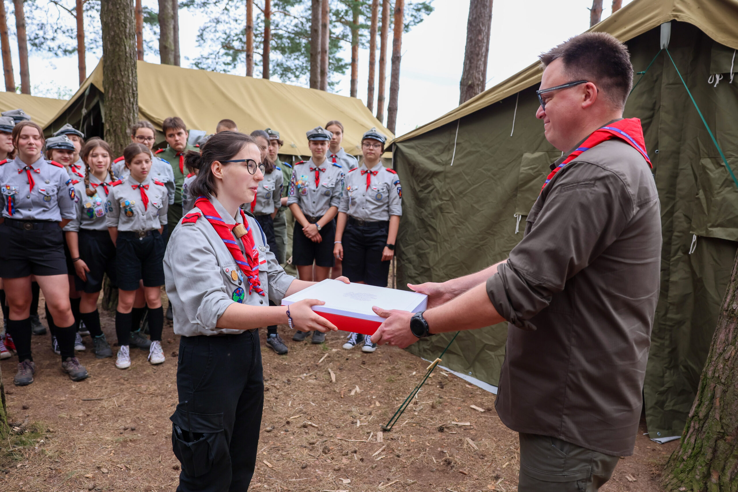 Marszałek Sejmu Szymon Hołownia wręcza Flagę RP harcerce podczas uroczystości w obozie harcerskim, otoczony przez innych harcerzy i namioty w tle.