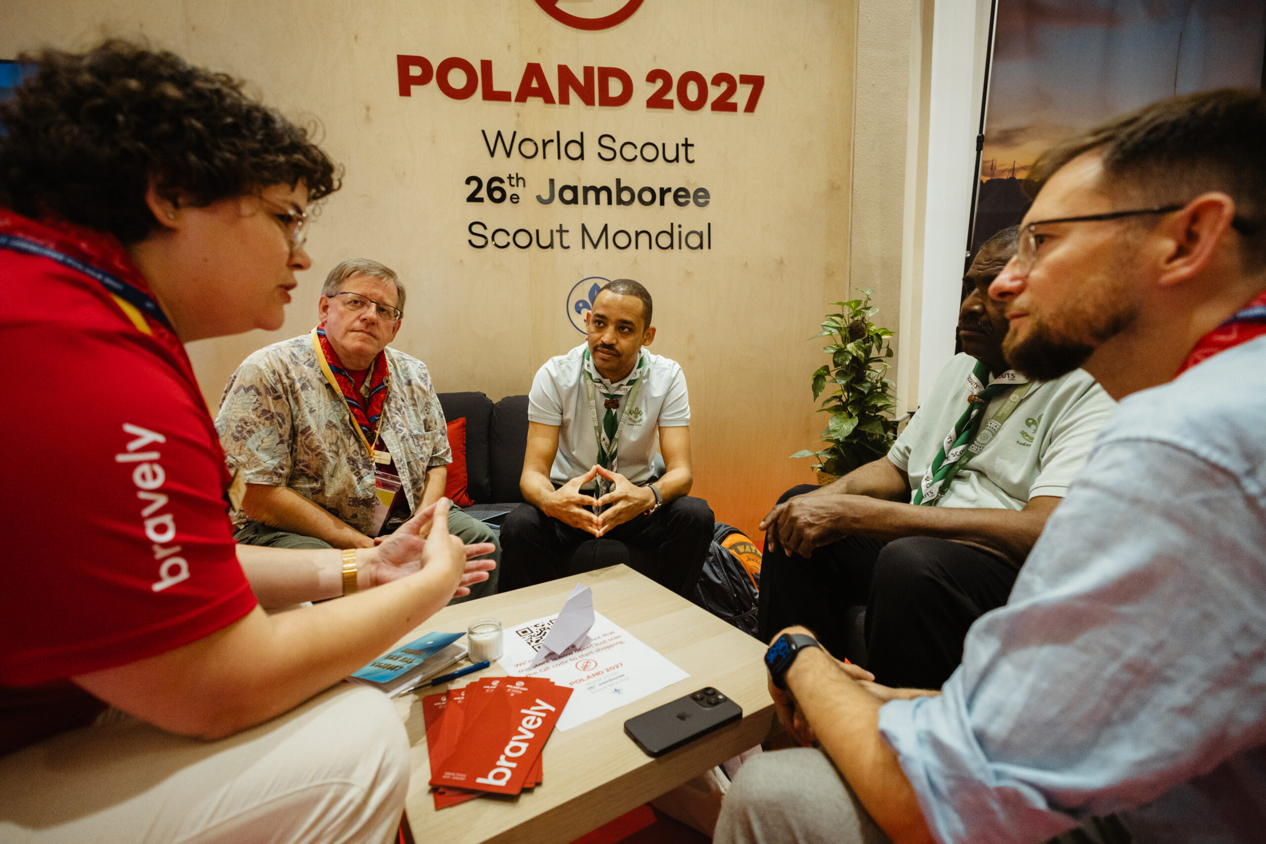 ZHP na 43. Światowej Konferencji WOSM w Egipcie! 1 Na obrazku widnieją 4 osoby, które siedzą przy niskim stole, na stole znajdują się czerwone ulotki "bravely". Z tyłu ścianka z napisem "Poland 2027".