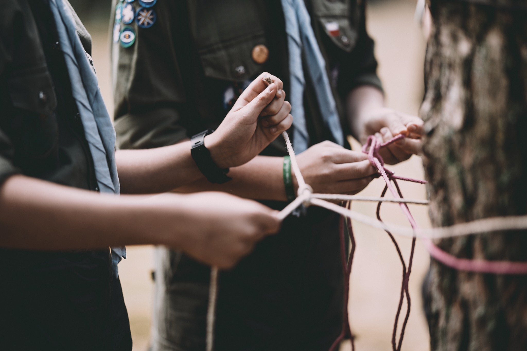 Inkluzywnośc w ZHP - harcerstwo dla dzieci z niepełnosprawnościami, na zdjęciu widać dłonie harcerzy, którzy wspólnie uczą się wiązać węzły. W tel widać rozmyte mundury ze sprawnościami.