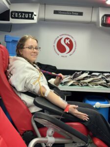 Harcerska służba, która ratuje życie. Krwiodawstwo w ZHP 2 Harcerska służba - krwiodawstwo w ZHP. Dziewczyna w białej bluzie w trakcie oddawania krwi, siedzi na czerwonym fotelu, pokazuje na harcerską chustę zawieszoną na szyi.