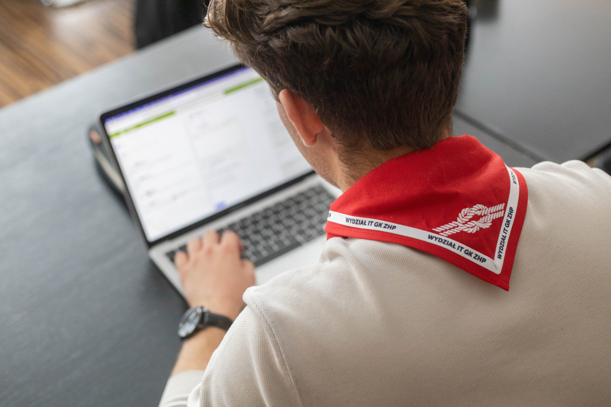 Wydział IT w ZHP - wsparcie dla organizacji - mężczyzna z czerwoną harcerską chustą z nazwą wydziału, patrzy w komputer i pracuje.
