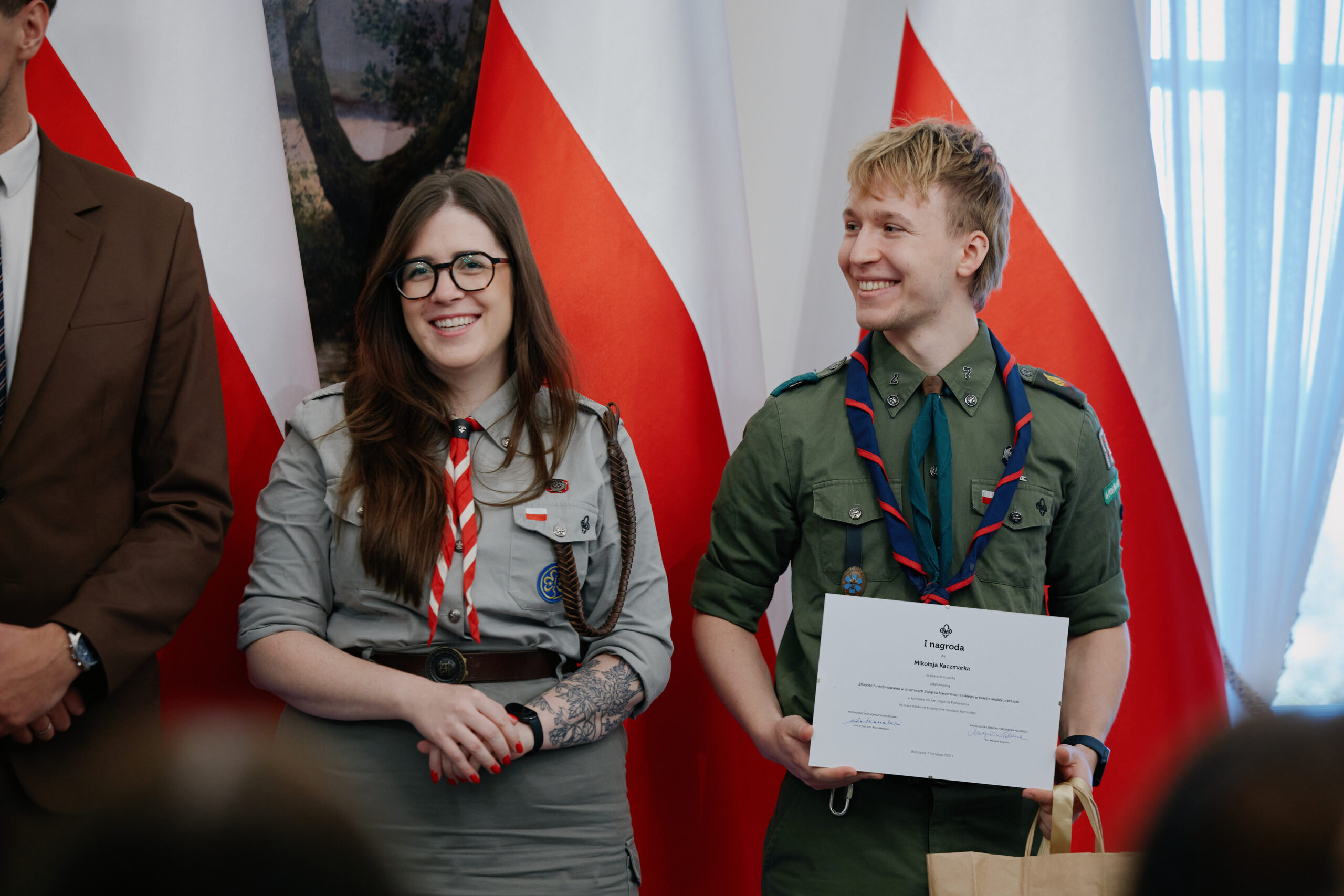 Naczelniczka ZHP oraz harcerz podczas uroczystości wręczenia nagrody za zajęcie miejsca w konkursie na prace naukowe o tematyce harcerskiej, stoją na tle biało-czerwonych flag; laureat trzyma dyplom i uśmiecha się.