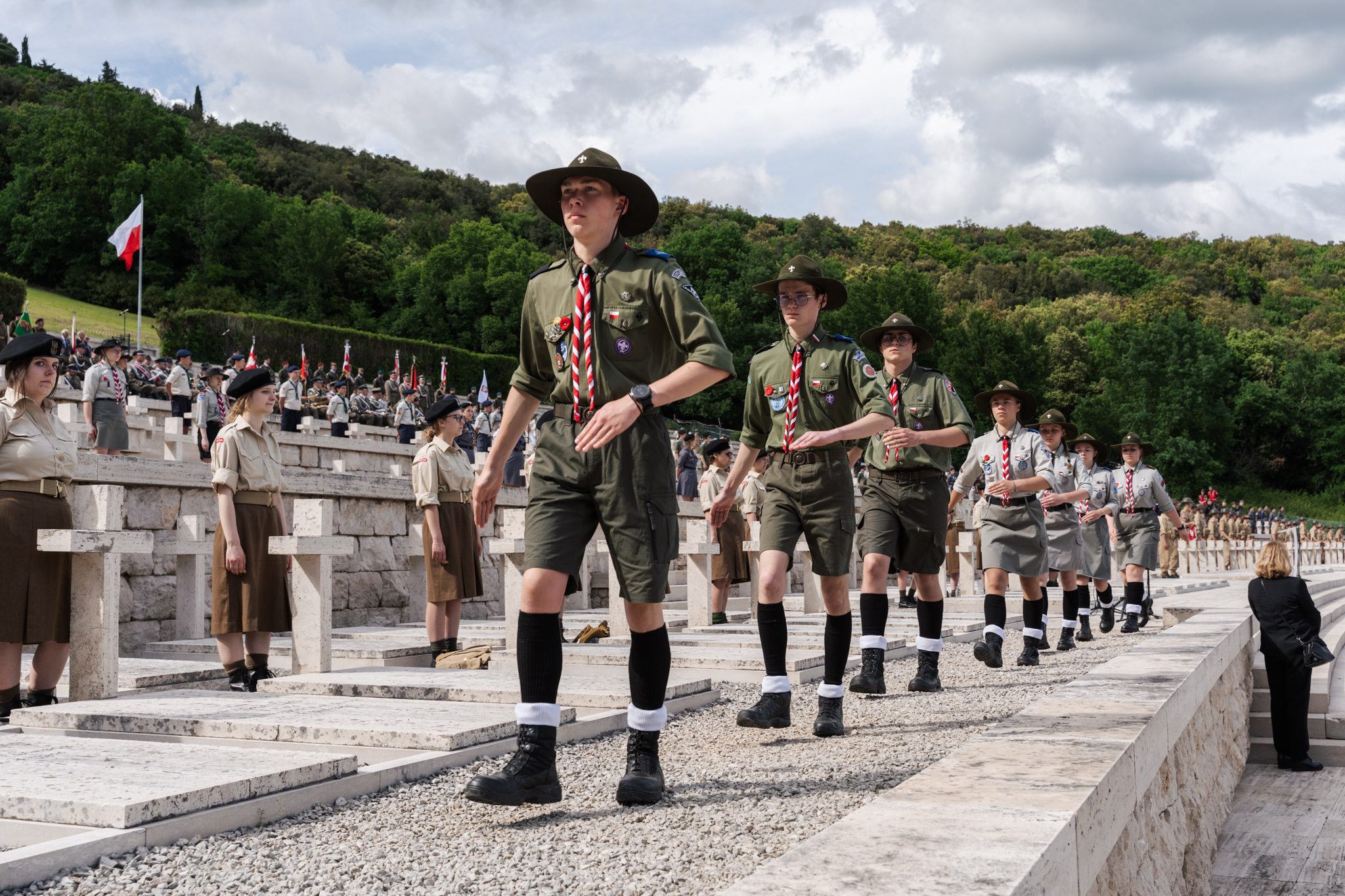 Wyprawa pamięci - harcerze i harcerki w mundurach maszerują w rzędzie wzdłuż aleji cmentarza na Monte Cassino.